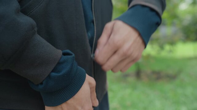 Person fastening coat outdoors, Close view of hands securing coat in scenic park setting, Detailed shot of person zipping jacket with textured fabric under gentle sunlight in park environment