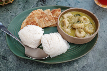 A green plate serving a meal of Thai green curry with fish balls, accompanied by crispy roti flatbread and bundles of white rice noodles.