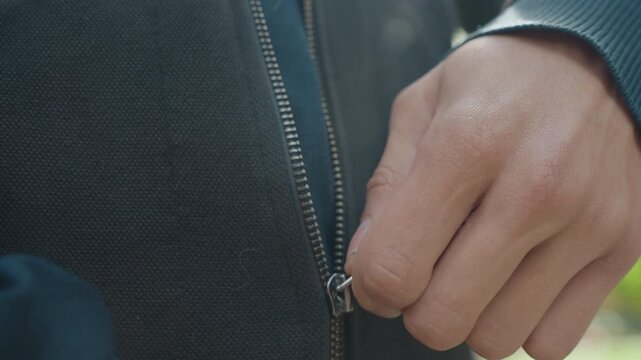 closeup hands fastening zipper on jacket, caucasian man outdoors in park adjusting hoodie during morning routine, steady fingers working slider, textured fabric and metal teeth visible, soft sunlight
