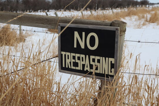 A black and yellow no trespassing sign, nailed to a fence post. The sign is obscured by dead grass poking through a layer of snow.