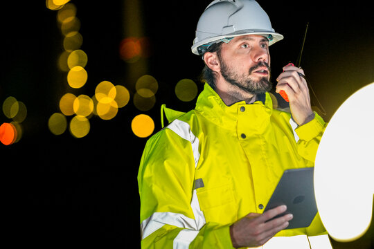 Safety officer reporting urgent situation via walkie talkie at night. Industrial worker in high-vis jacket using tablet to check accident risk data onsite.