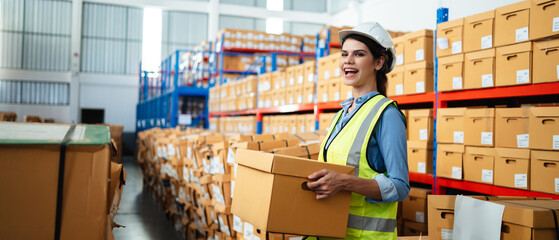 Professional female warehouse worker in safety vest and hard hat holding a cardboard box in a large storage facility. Logistics, inventory management, and industrial supply chain concept.