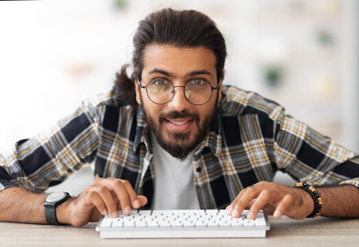 Young middle-eastern man programmer sitting at worktable, using modern miniature pc keyboard, wearing glasses, closeup portrait. Happy arab guy developer working from home, enjoying his job