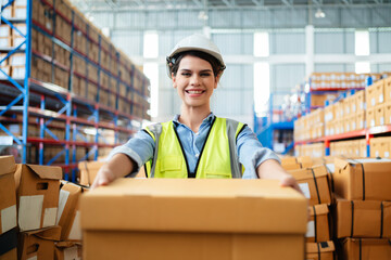 Professional female warehouse worker in safety vest and hard hat holding a cardboard box in a large storage facility. Logistics, inventory management, and industrial supply chain concept.