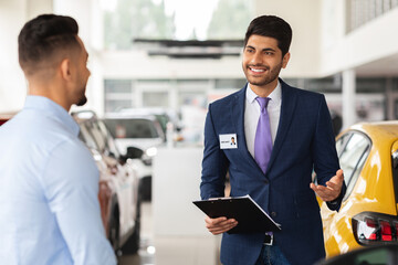 Cheerful VIP car salon manager having conversation with male customer, giving useful tips for choosing automobile, two handsome middle-eastern men standing among nice brand new autos