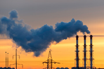 Dark factory smoke drifts across orange sunset sky above power lines and shows industrial emissions, energy demand, climate risk.