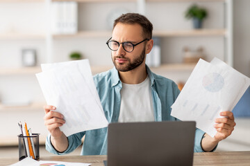 Serious young man looking through documents while having online business meeting at home office. Focused male freelancer making web presentation, communicating remotely on laptop