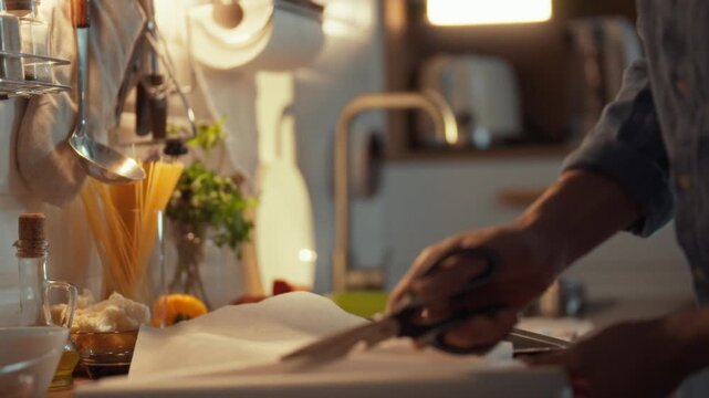 A man carefully cuts parchment paper with kitchen scissors to perfectly fit a baking tray, preparing to lay out culinary delights in the kitchen for a precise bake