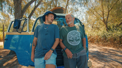 Two relaxed men stand near the retro bus loaded with surfing boards and watch somewhere. Modern hippies travel by retro bus and check waves on a surfing spot