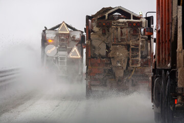 Convoy of road service salt spreader trucks moves through heavy blizzard on winter highway, flashing beacons cut foggy spray.