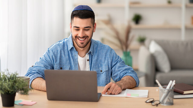 Telecommuting Concept. Potrait of smiling casual israeli man sitting at desk, using laptop. Male watching webinar looking at pc screen, working or studying remotely, free copy space, banner