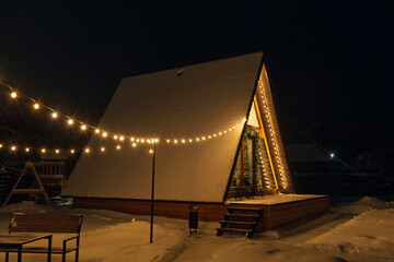 Cozy A-frame wooden cabin decorated with warm lights at night in snowy landscape, creating calm winter holiday mood and outdoor retreat atmosphere.