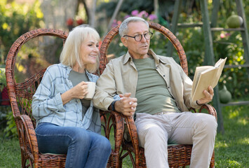 Loving elderly couple reading book together, sitting in wicker chairs and spending warm spring evening outdoors in their garden. Woman drinking coffee and spouses holding hands