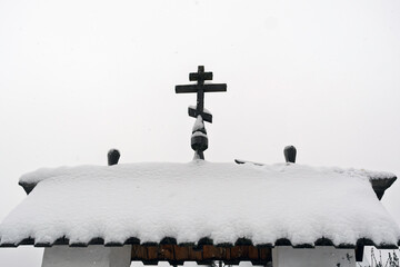 Snow-covered wooden roof with traditional cross against white winter sky, minimal rural architecture scene with calm cold atmosphere.