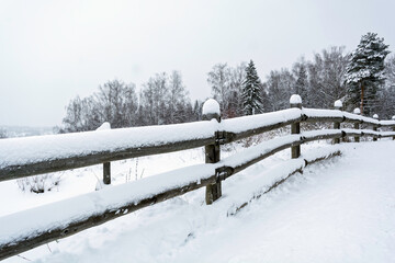 Snow-covered wooden fence along winter path with bare trees, quiet countryside landscape showing cold weather and rural calm.