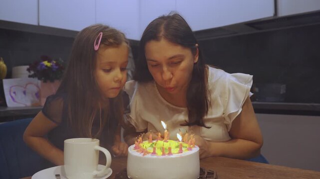 A mother blows out the candles on a cake during a small, homey celebration of her birthday with her daughter in the kitchen. Celebrating a birthday at home. Mother and daughter, happy parenting.