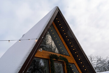 Triangular A-frame house facade with snow-covered roof and glowing lights seen from below, creating warm contrast against cold winter sky.