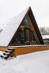 Snow-covered A-frame wooden cabin with terrace and outdoor chairs stands in quiet winter yard, decorated with lights for seasonal rest.