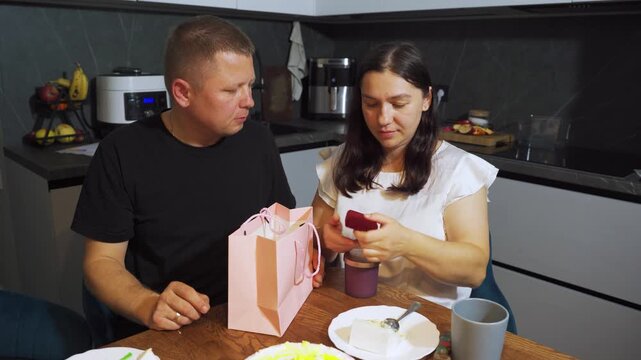A woman receives a gift from her husband in the kitchen and rejoices. Family relationships, family evening, a man congratulates his wife in the kitchen and gives her a gift.