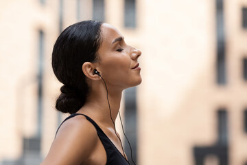 Glad young arab female with closed eyes in headphones enjoy music and relaxing on building background outdoor, profile, close up. Sports on street with device, weight loss, body care and training