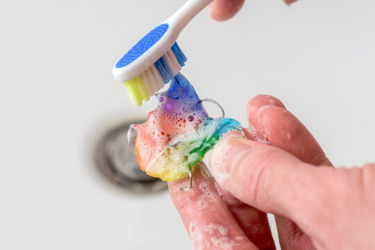 Adult cleans child orthodontic retainer above sink with toothbrush and soap foam, daily routine helps safe oral appliance care.