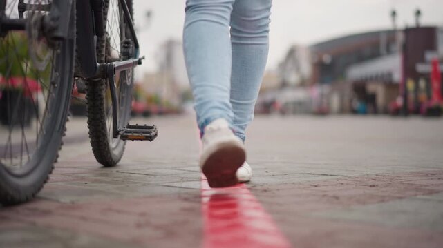 caucasian woman walking red line pavement, bicycle parked, closeup on sneakers and jeans, slow measured steps, storefronts blurred, damp cobblestone texture, soft overcast light, solitary commuter