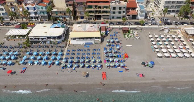 Aerial view of beach clubs featuring many colorful parasols and sunbeds arranged on a grey pebble shore. The clear turquoise water borders the beach. It is located in Letojanni beach, Sicily, Italy.