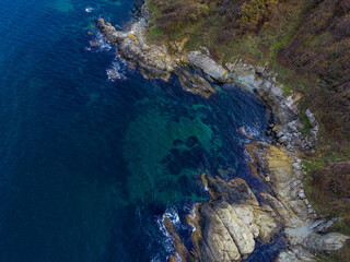 Waves crash against rocks along the coastline near a clear blue sea on a sunny day with some greenery around