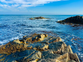 Waves crash on rocks along the shoreline under a clear blue sky during the day near the coast