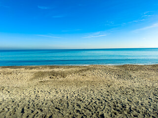 Bright blue ocean meets sandy beach under a clear sky during midday near the coast, showing still water and wide shoreline