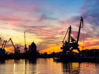 Bright colors fill the sky as cranes stand tall in a busy harbor at sunset while small boats float on calm water below