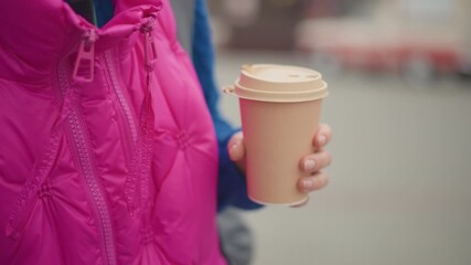 woman holding takeaway coffee cup closeup, pink puffer jacket with zipper detail, single hand gripping warm paper cup, blurred city street background, cool weather commute vibe, casual fashion