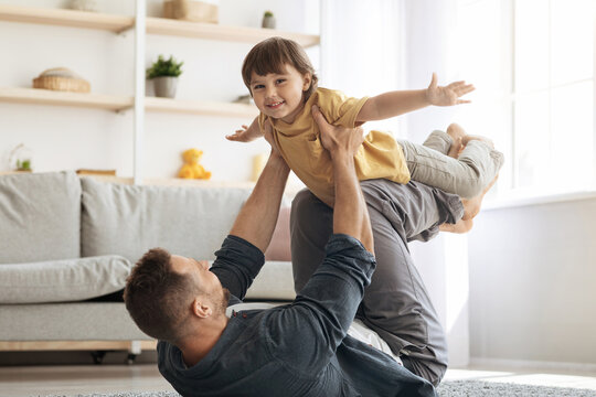 Happy young caucasian father lying on floor and carrying his little son on hands, kid playing plane and smiling to camera, funny domestic activities concept