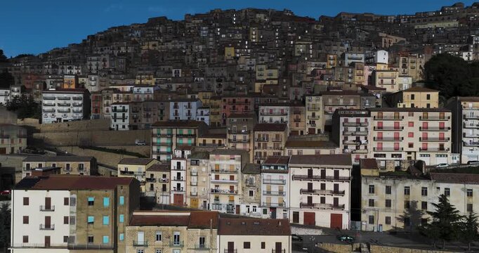 Aerial view of closely stacked buildings of varying heights and colors clinging to a hillside under a clear sky, Gangi, Sicily, Italy.