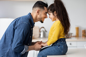 Obraz premium Father Child Love. Happy Middle Eastern Dad Bonding With Little Daughter At Home, Cheerful Arab Daddy Holding Hands And Touching Foreheads With Cute Female Kid That Sit On Table, Side View