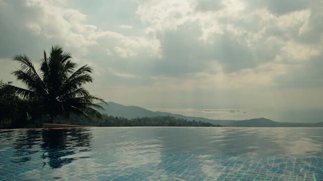 Infinity pool with calm water reflecting cloudy sky and distant tropical hills, serene panoramic landscape with palm tree silhouette