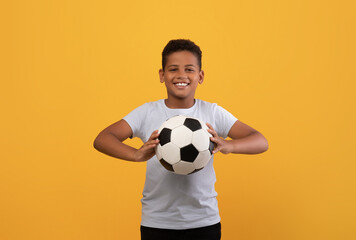 Cheerful black school boy holding soccer ball and smiling at camera, playing football, yellow studio background, copy space. Sport, leisure, entertainment, hobby for kids concept