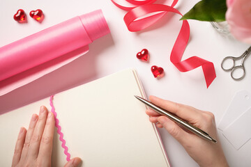 Hands writing in blank notebook surrounded by pink wrapping paper, red hearts, and ribbon. Romantic planning with copy cpace
