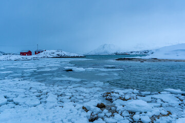 Red wooden houses stand along a frozen coastal shoreline with drifting ice and snow-covered mountains in the background. The quiet winter scene captures the cold, remote atmosphere of a Nordic or Arct © murattellioglu