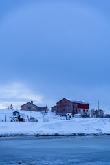 Red wooden houses stand along a frozen coastal shoreline with drifting ice and snow-covered mountains in the background. The quiet winter scene captures the cold, remote atmosphere of a Nordic or Arct © murattellioglu
