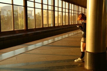 Teen girl waiting in a subway station with windows in sunlight. Young woman standing against a column in style clothes, looking in her mobile phone, natural person pose. Indoors perspective concept