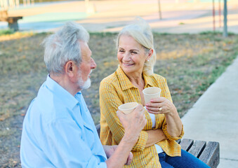Happy active senior couple having fun talking drinking coffee and bonding sitting on bech in park...