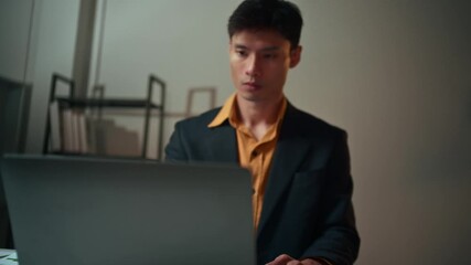 Focused asian businessman using a laptop and calculator while working late. Overworked employee analyzing financial data and charts at his desk - Powered by Adobe