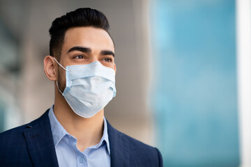 Middle-eastern guy businessman in medical face mask and formal outfit standing by office building, looking at copy space, closeup portrait. Business while COVID-19 pandemic concept