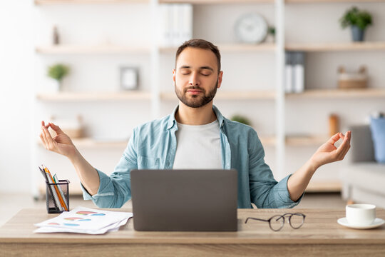 Workplace stress management. Calm Caucasian man meditating with closed eyes in front of laptop pc at home office. Young male freelancer feeling peaceful and balanced, doing yoga at his desk