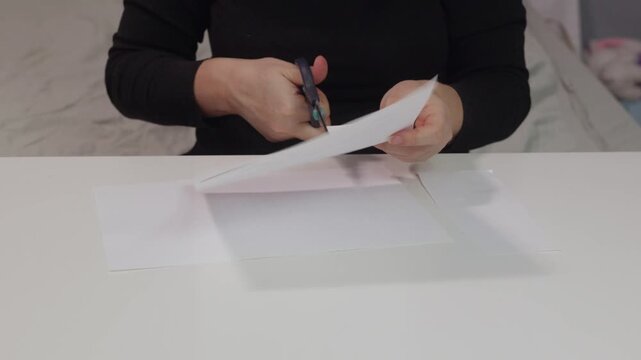 Close up of adult woman hands cutting white paper sheet with scissors on table indoors, illustrating manual craft, preparation and creative work concept.