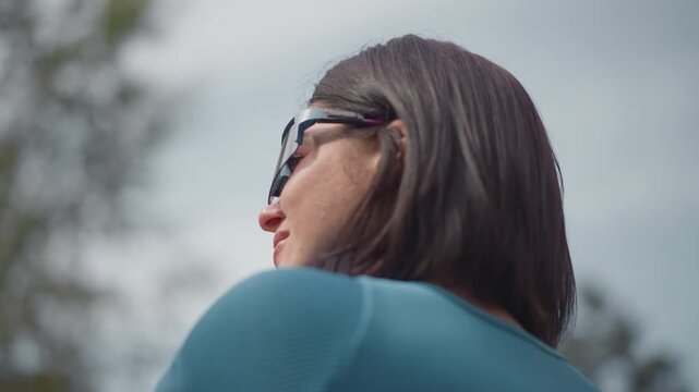 woman adjusting bicycle helmet outdoors, back profile showing bob hairstyle and teal jersey, fastening strap and visor, overcast sky with blurred trees, calm focused mood, commuter preparing for solo