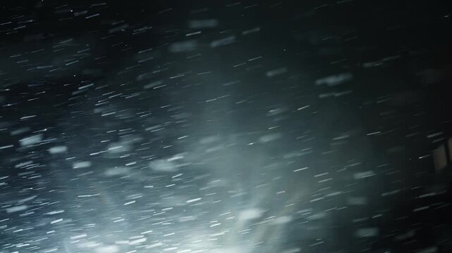Backlit snowflakes streaking across the dark night sky during a gusty winter storm, illuminated by a distant light source and capturing the dynamic motion of falling snow.