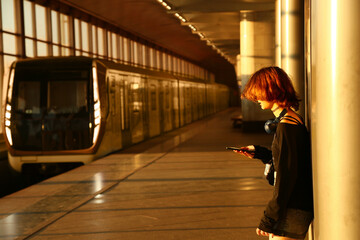 Teen girl waiting in a subway station with windows in sunlight. Young woman standing against a column in style clothes, looking in her mobile phone, natural person pose. Indoors perspective concept