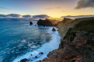 Dramatic ocean coastline at sunset with rugged cliffs, powerful waves and golden light breaking through clouds. The seascape captures the raw beauty of nature, where deep blue water meets dark rocky.
